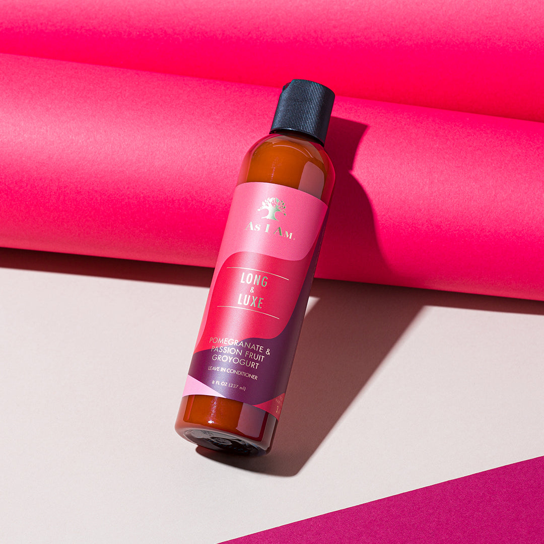 En brun flaske As I Am Long & Luxe GroYogurt Leave-in Conditioner (237 ml) med en lyserød etiket, der ligger diagonalt foran en lyserød baggrund.