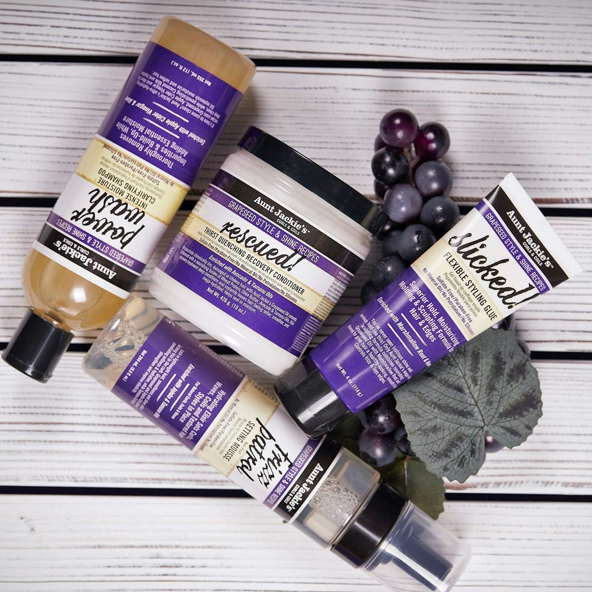 Udvalg af Aunt Jackie's hårprodukter: Power Wash Shampoo, Rescued Conditioner, Slicked Styling Glue og Frizz Patrol Mousse, ledsaget af mørke druer.