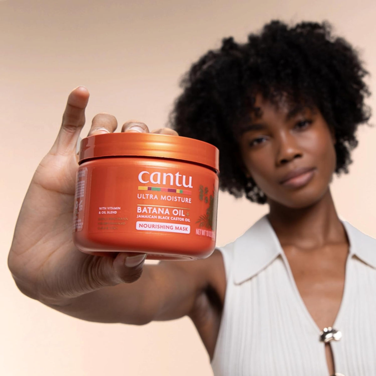 Kvinde med naturligt krøllet hår holder Cantu Ultra Moisture Batana Oil nærende hårmaske frem mod kameraet.